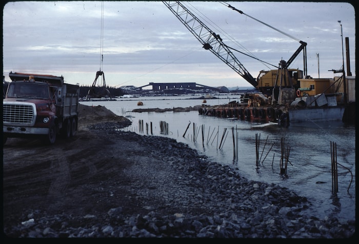Photographie d'archive d'un chantier de construction portuaire en bordure d'eau. Sur la gauche, un camion-benne est stationné sur une rive recouverte de pierres concassées. Des tiges de métal verticales sortent de l'eau le long de la berge.