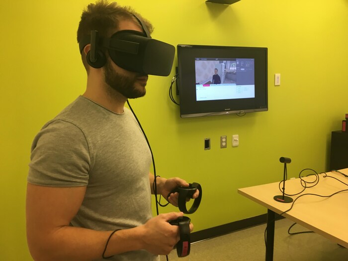 On voit un homme muni d'un casque de réalité virtuelle et de manettes reliées par des fils. Sur le mur, un écran affiche l'image de réalité virtuelle : le patient dans sa maison. 