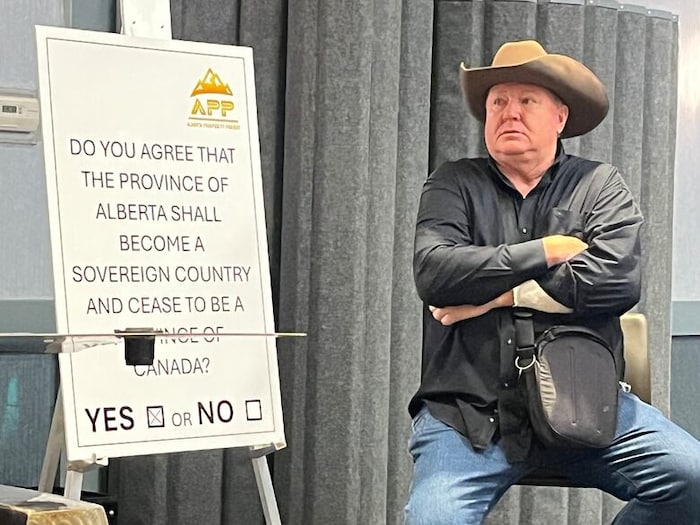 Jeff Rath est assis sur un tabouret à côté d'une affiche qui indique une question référendaire sur la souveraineté de l'Alberta. 