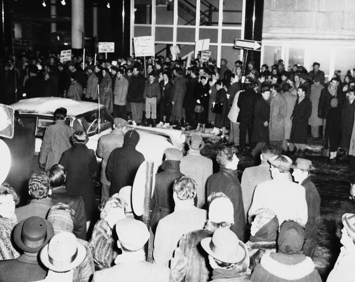 Lors de la grève des réalisateurs de Radio-Canada en 1958-1959, à l'extérieur à
5h30 PM à proximité de l'édifice de Radio-Canada, une voiture circule sur une
rue où une foule compacte est massée de chaque côté sur les trottoirs. Parmi les gens présents, des grévistes avec des pancartes.