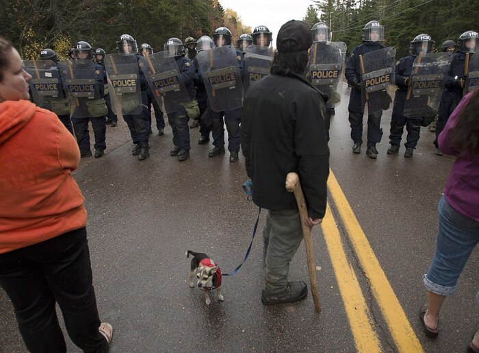 Rangée de policiers antiémeute devant un manifestant avec une béquille et un très petit chien.