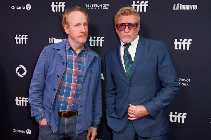 Deux hommes posent pour une photo. Celui de droite est en complet bleu et porte des lunettes fumées roses. Celui de gauche a une chemise carreautée, des jeans et une veste en jean.