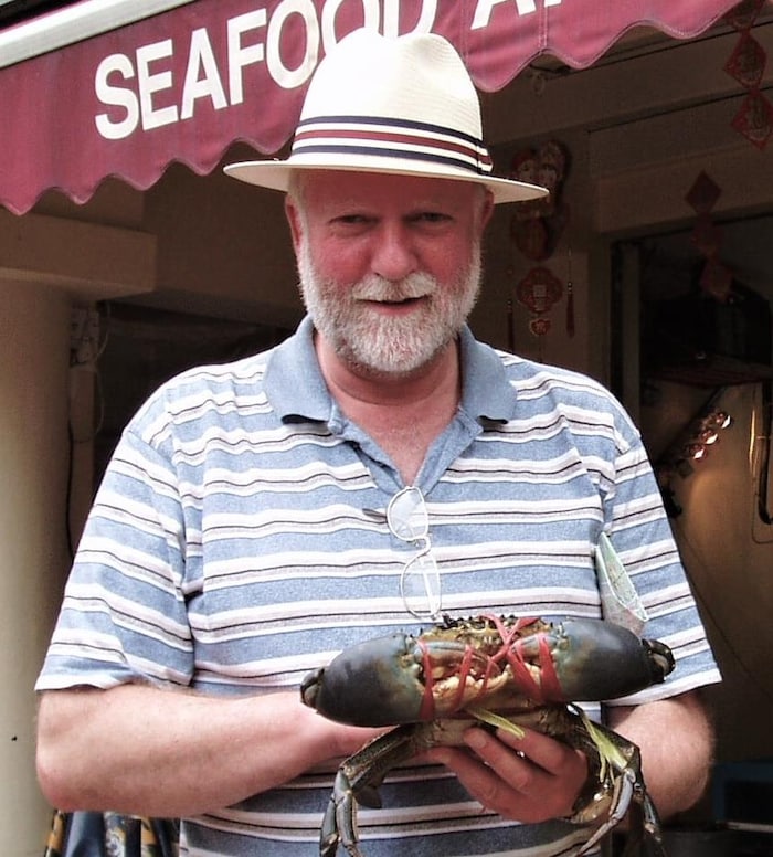 Un homme portant une courte barbe blanche et un chapeau d'été blanc est debout devant un marché de fruits de mer et tient un crabe vivant aux pinces retenues par des élastiques.