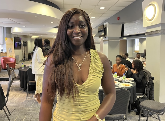 Une femme en blouse jaune pose pour une photo, debout dans une salle de conférence avec des personnes en arrière-plan assises à des tables rondes ou debout.