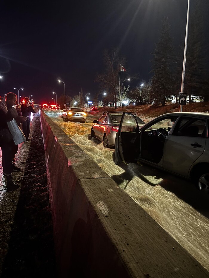 Plusieurs véhicules sont immobilisés dans un important courant d'eau boueuse qui inonde une autoroute la nuit. Une portière de voiture est ouverte au premier plan, tandis que des témoins observent la scène depuis le bord de la route derrière une murette de béton.