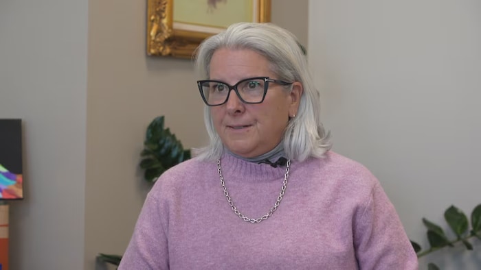 Une femme portant des lunettes et un pendentif au cou fixe du regard l'objectif d'une caméra.