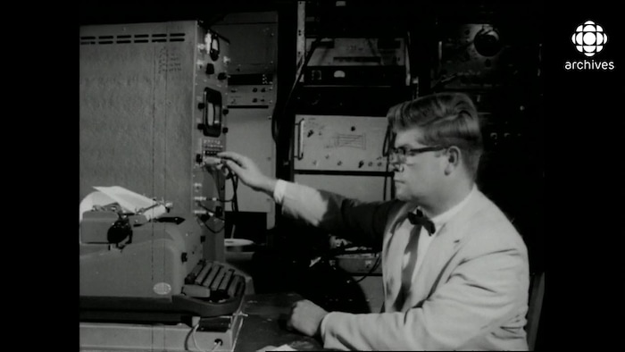 Homme bien mis et bien vêtu en veston avec noeud papillon devant une machine à écrire tout en manipulant des câbles sur un moniteur