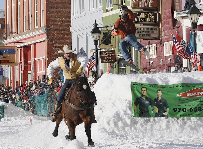 Devant des enseignes sur des maisons pittoresques, un skieur bondit au-dessus d'un gros banc de neige en tenant une corde attachée à un cheval chevauché par un cow-boy.