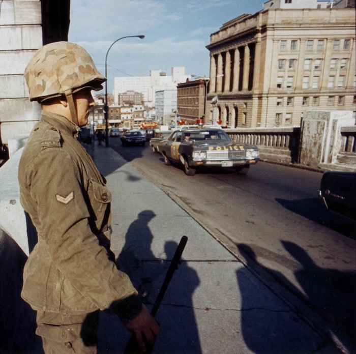 Un soldat des Forces canadiennes, vu de profil, assure la garde à l'entrée d'un pont sur lequel s'engage une voiture de police. 