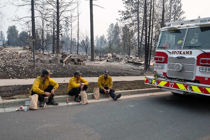 Trois pompiers assis à côté de leur camion, devant des maisons en ruine.