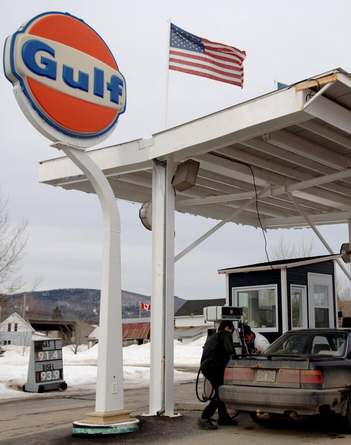Deux hommes autour d'une auto très sale, immatriculée au Québec, pendant un plein d'essence à une station-service Gulf sur laquelle flotte un drapeau des États-Unis.