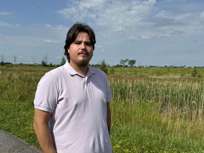 Un homme vêtu d’un polo clair pose devant un vaste terrain couvert d’herbes hautes, sous un ciel nuageux.