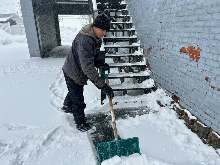 Sous la neige, un homme avec une pelle gratte quelques centimètres de neige en bas d'un escalier.