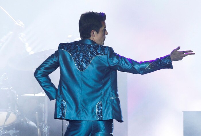 Brandon Flowers de dos, portant une veste bleue et brillante, d'inspiration country, et pointant vers sa droite avec l'index et le majeur.