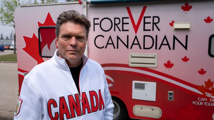 Un homme portant un tricot sur lequel il est écrit CANADA en lettres capitales sur un fond rouge se photographie devant un autocar habillé en rouge et portant un écriteau en anglais Forever Canadian.