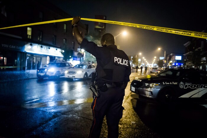 Un policier en uniforme soulève un ruban, qui sert à fermer un secteur lorsque les policiers érigent un périmètre de sécurité. La photographie a été prise le soir, car il fait sombre et seuls les néons et lampadaires illuminent la scène. 