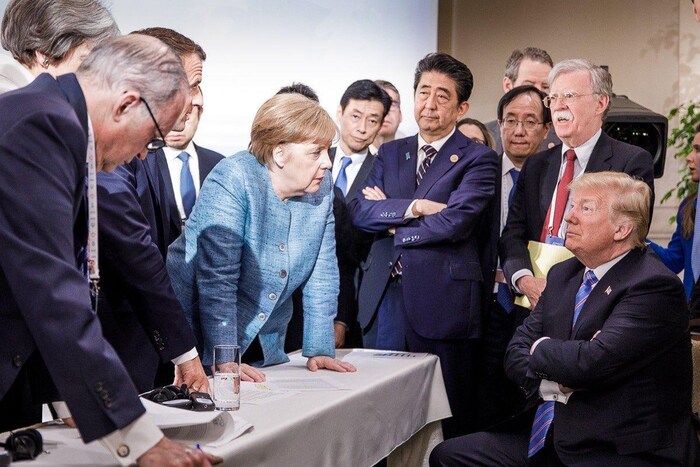 Donald Trump, assis, semble se faire gronder par Angela Merkel, debout devant lui. 