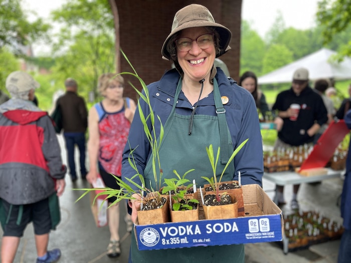 Une femme sourit à belles dents en tenant plusieurs plantes dans une boîte en carton de vodka soda.