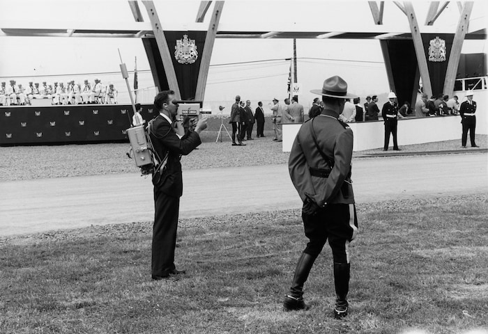 Un caméraman de Radio-Canada, portant son équipement sur son dos et des membres de la Gendarmerie royale du Canada sur le site de la cérémonie d'inauguration de la Voie maritime du Saint-Laurent.