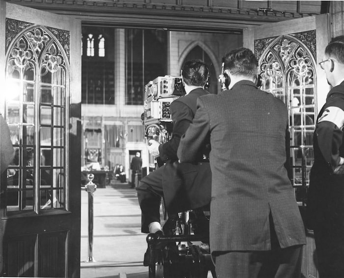 Un caméraman équipé d'une caméra sur un chariot roulant entre au parlement accompagné de deux techniciens de Radio-Canada.