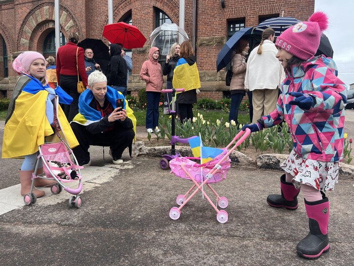 Lors d'un rassemblement en plein air, une femme agenouillée, un drapeau ukrainien autour des épaules, prend une photo d'une petite fille tenant une petite poussette rose décorée de deux petits drapeaux de l'Ukraine.