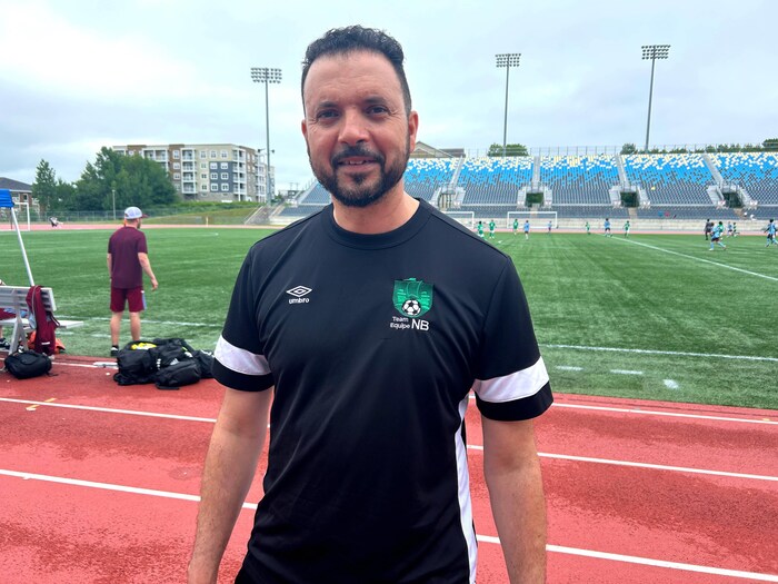 Un adulte vêtu d'un chandail de Soccer NB pose dehors pour une photo, debout sur la piste d'athlétisme qui entoure le terrain de football.