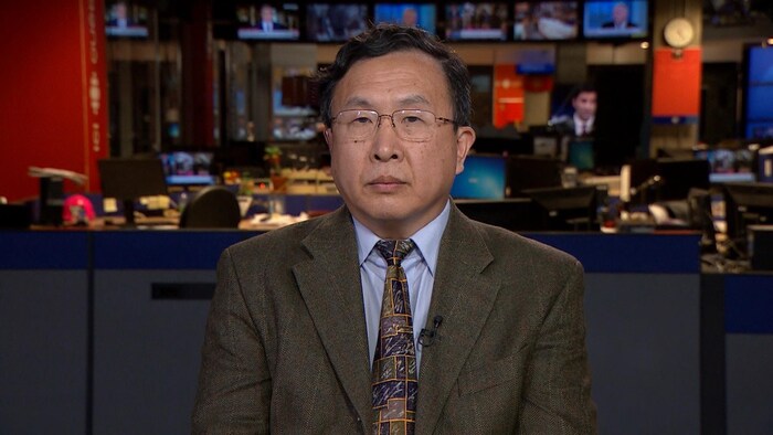 Un homme asiatique portant des lunettes, un veston et une cravate, regarde la caméra sans sourire.