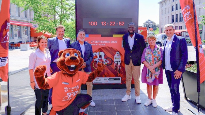 Six personnes et une mascotte posent devant une horloge numérique.