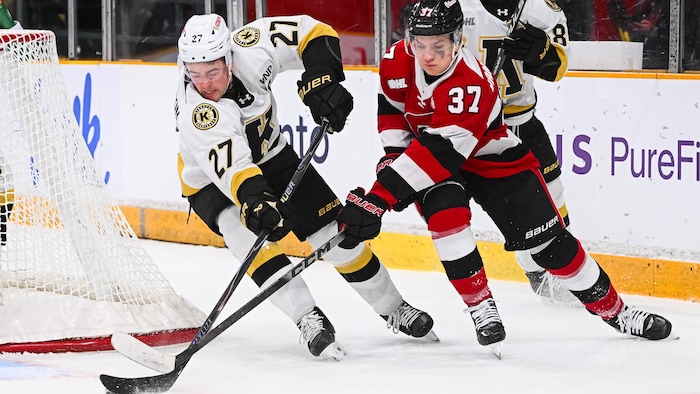 Un joueur de hockey habillé en blanc, rouge et noir tente d'enlever la rondelle à son adversaire pendant un match junior.