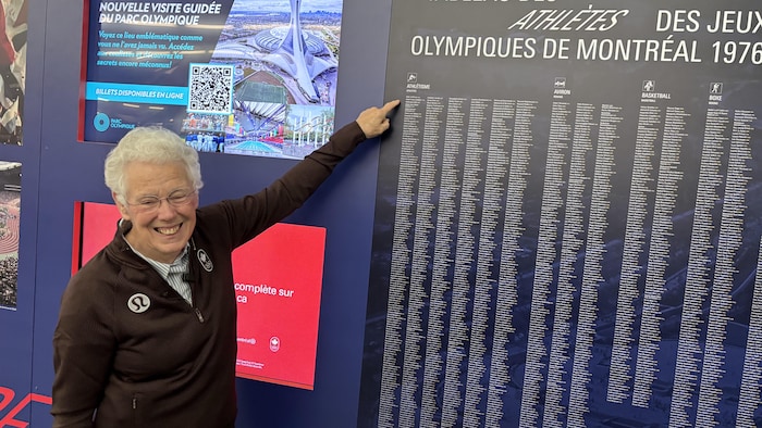 Elle sourit en pointant un écriteau qui montre la liste des athlètes des Jeux de Montréal.