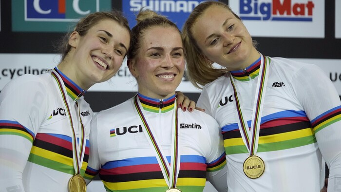 Trois cyclistes sourient sur le podium, chacune avec une médaille d'or au cou. 