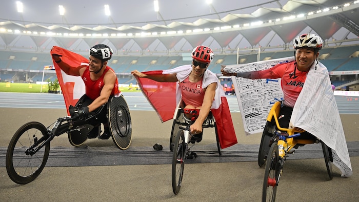 Trois athlètes en fauteuil roulant sourient avec le drapeau de leur pays respectif autour de la taille.