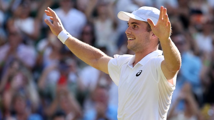 Un joueur de tennis en blanc sourit en saluant la foule qui l'acclame. 
