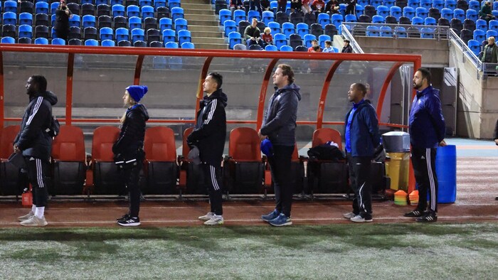 Personnes debout, de profil, devant un banc de joueurs.
