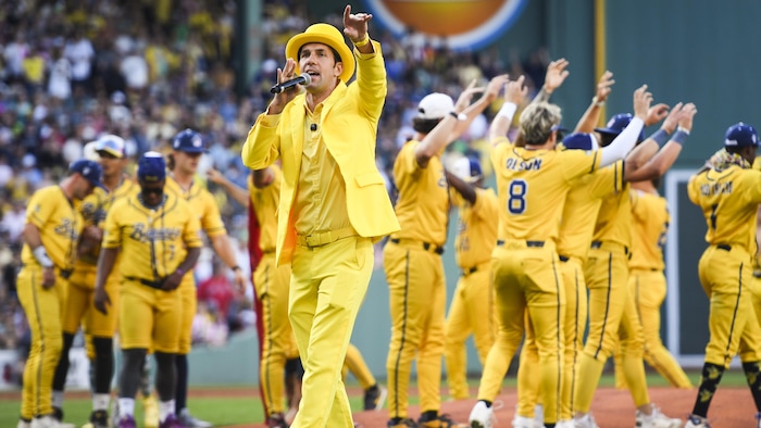 Un homme sur un terrain de baseball parle en tenant un micro devant des joueurs.