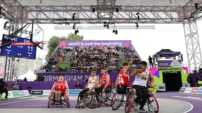 Deux équipes s'affrontent lors d'un match de basketball en fauteuil roulant, devant une tribune pleine, à l'extérieur. 