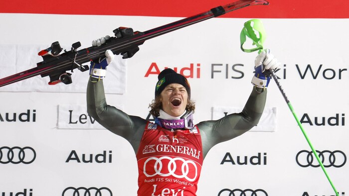 Il crie sur le podium et tient ses skis bien haut dans sa main droite et un bâton dans sa main gauche.