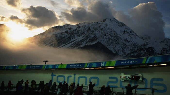 Un bobsleigh biplace glisse sur une piste. Il est écrit Torino 2006 dans la glace et on voit une montagne en arrière-plan.  