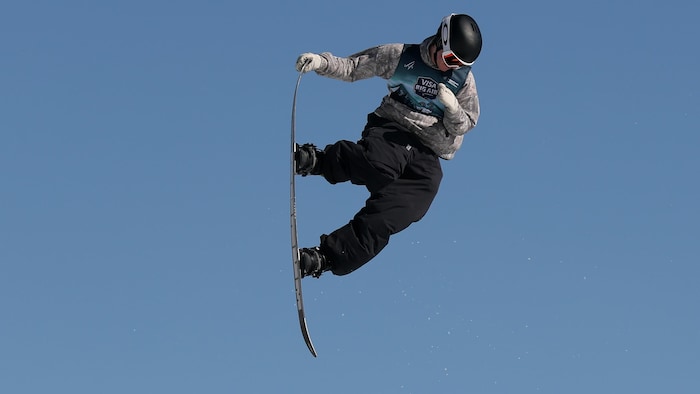 Éli Bouchard, dans les airs, tient le bout de sa planche de la main droite. 