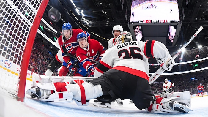 Deux joueurs de hockey devant un gardien qui tente de faire un arrêt en étirant la jambière. 