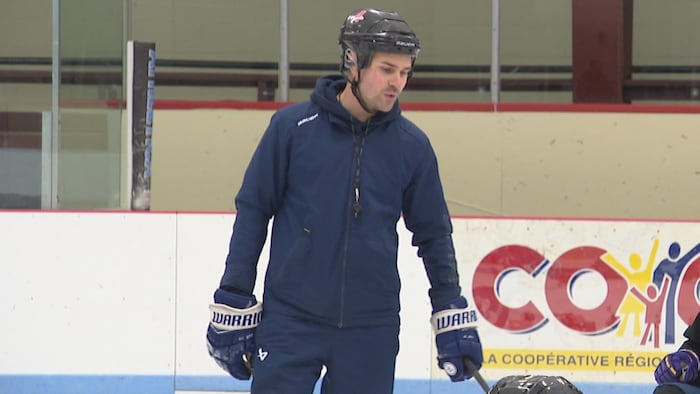 Carl Devost est sur la patinoire. Il porte un casque de hockey et s'adresse à des jeunes à l'extérieur du cadre de la photo.