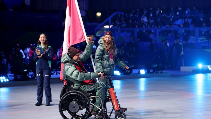 Mark Ideson, dans son fauteuil roulant, lève le poing au ciel et sourit. Un drapeau canadien est accroché à son fauteuil. À côté de lui, Natalie Wilkie marche et le regarde en souriant.