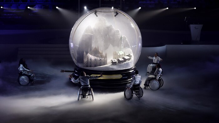 Cinq danseurs en fauteuil roulant encerclent une boule à neige géante qui contient un paysage rappelant les différents lieux des Jeux paralympiques de Milan-Cortina.