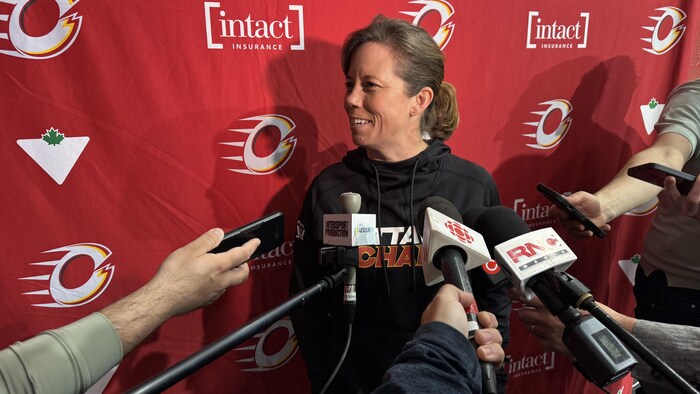 Une entraîneuse de hockey est en mêlée de presse avec des journalistes. On voit le logo de son équipe derrière elle.