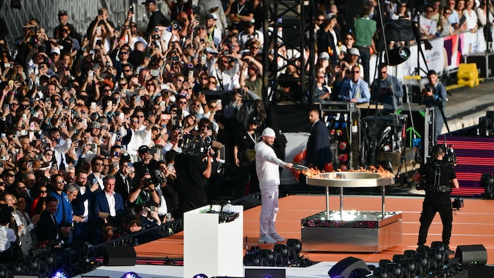 Un homme allume un grand rond d'aluminium avec la flamme olympique sur une scène, et des centaines de personnes le regardent.