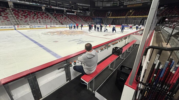 Homme assis sur le banc, qui regarde un entraînement sur la patinoire.
