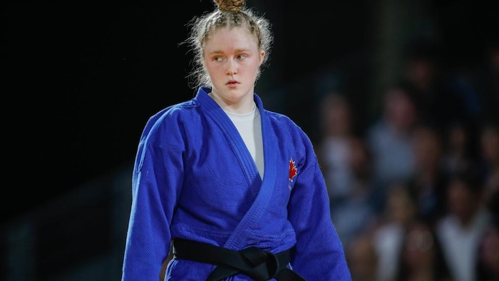 Une jeune femme dans un judogi, avec une grosse ceinture noire autour de la taille, se tient debout et regarde sur sa droite.  