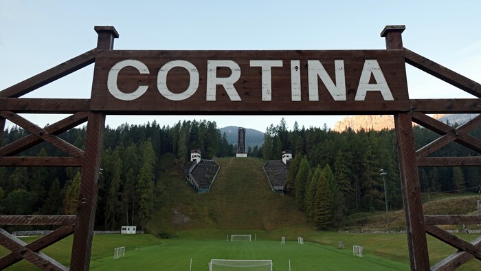 Photo d'un ancien site olympique l'été. À l'avant-plan une pancarte où il est inscrit : Cortina.