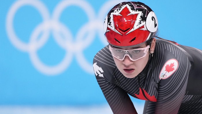 L'athlète est penchée sur une patinoire, lunette de sport sur le nez, devant les anneaux olympiques.