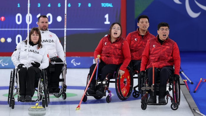 Pendant la finale de curling en fauteuil roulant, deux athlètes canadiens et trois athlètes chinois regardent une pierre se diriger vers la maison.